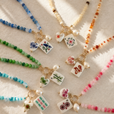 Six beaded necklaces with gold-tone hardware and white enamel game-piece charms are arranged on a textured cream fabric. The necklaces feature blue, cream, green, aqua, orange, and pink beads, each with a different painted tile charm including a bird, flowers, bamboo, cards, and symbols.