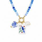 A beaded necklace with light blue and royal blue round beads, featuring a gold-tone clasp and three charms: a blue and white floral heart-shaped tile, a white playing card charm with a blue bird and red flowers, and a white pearl bead.