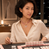 A woman in a white blouse sits at a mahjong table, holding a tile and wearing a pink beaded necklace with a dangling pendant. Mahjong tiles are arranged on the table in front of her.
