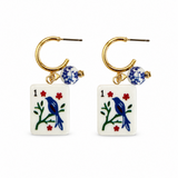 A pair of gold hoop earrings with rectangular white playing card tiles featuring blue birds and red flowers, and a small blue-and-white patterned bead dangle, shown on a plain white background.