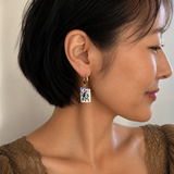 A close-up side profile of a woman with short black hair wearing a gold hoop earring with a rectangular white enamel charm decorated with blue and green floral details.