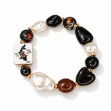 A beaded stretch bracelet featuring a white square tile charm with a black and orange koi fish design, alternating with glossy black stones, marbled black-and-orange beads, white baroque pearls, and a smooth brown stone, all separated by gold spacer rings on a white background.