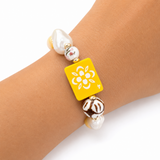 Bracelet with yellow square charm featuring a white flower design on a wrist.