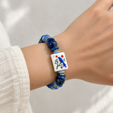 Blue mahjong tile bracelet worn on a wrist, featuring navy beads, blue stone chips, gold-tone spacers, and a white tile with a blue bird and red flowers.