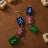 A pair of dangling earrings designed to look like mahjong tiles, featuring blue, pink, and green tiles with white symbols, hanging on gold-tone hooks on a wooden tabletop surrounded by real mahjong tiles.