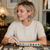 A woman with short blonde hair wearing a cream knit sweater and colorful geometric drop earrings is playing a game of mahjong in a warmly lit indoor setting.