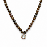 A beaded necklace featuring round brown and black tiger's eye beads with a small oval rose gold pendant set with a clear round gemstone, shown against a plain white background.