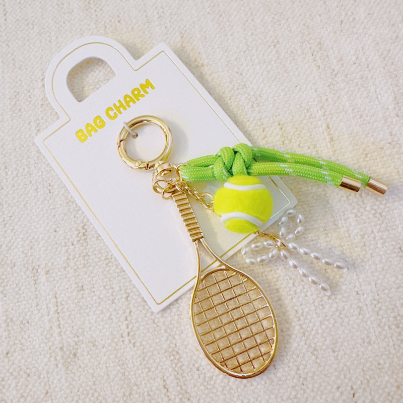 Gold-tone tennis-themed bag charm featuring a miniature racket, neon green tennis ball, and light green ribbon accents