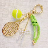 Gold-tone tennis-themed bag charm featuring a miniature racket, neon green tennis ball, and light green ribbon accents