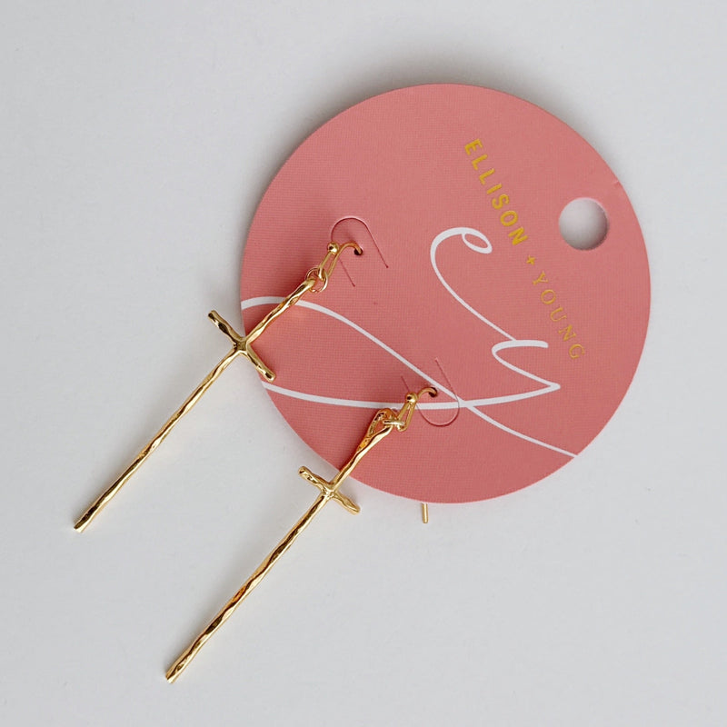 A pair of gold-tone molten-style cross earrings featuring a sleek, abstract design with a softly curved silhouette and minimal detailing, hanging vertically from simple hooks.