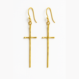 A pair of gold-tone molten-style cross earrings featuring a sleek, abstract design with a softly curved silhouette and minimal detailing, hanging vertically from simple hooks.