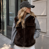 A woman wearing a dark brown faux-fur zip-front vest over a gray hoodie, with a black baseball cap, standing outdoors against a stone building wall.