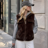 A woman wearing a dark brown faux fur zip-up vest over a gray hoodie and white pants, standing on a city sidewalk next to a stone wall.