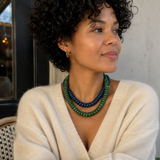 A woman with curly black hair wearing a cream knit sweater and a double-strand beaded necklace in green and dark blue, posed in a cafe setting.