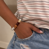 A close-up of a person's wrist wearing a beaded stretch bracelet with white, amber, and dark brown beads, paired with a striped t-shirt and denim jeans.
