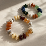Two beaded stretch bracelets with polished multicolor stones and small gold spacer beads, arranged on a light beige fabric background with soft diagonal shadows.