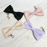 Four bow-shaped keychains in black, pink, beige, and purple on a light wooden surface.
