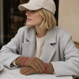 A woman seated at a table wearing a light gray wool coat, cream sweater, beige baseball cap, and tan knit gloves with bright pink trim.