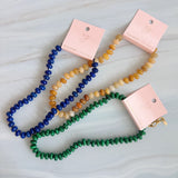 A collection of polished gemstone bead necklaces in various colors, including deep blue, green, brown, pink, and cream. Each necklace features smooth, rounded beads with a glossy finish, offering a timeless and versatile accessory look.