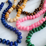 A collection of polished gemstone bead necklaces in various colors, including deep blue, green, brown, pink, and cream. Each necklace features smooth, rounded beads with a glossy finish, offering a timeless and versatile accessory look.