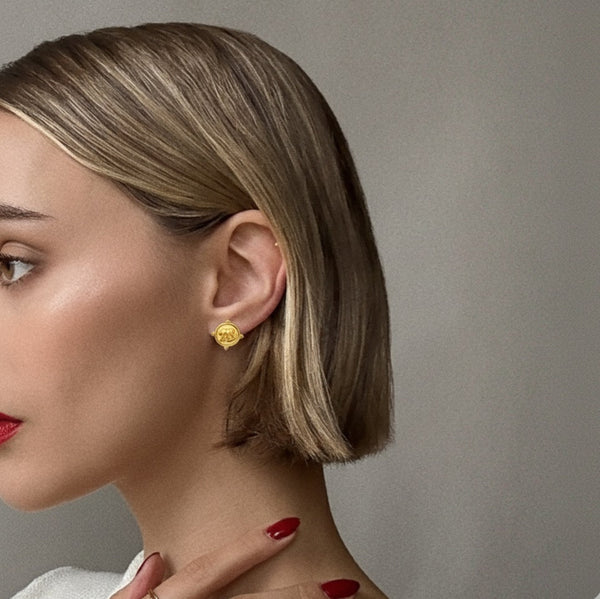 Close-up of a pair of round gold-tone stud earrings featuring a raised elephant motif on a medallion-style base with detailed edge accents.