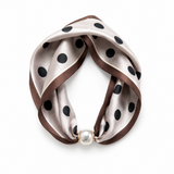 A twisted silk scarf with black polka dots on a champagne beige background, finished with a brown satin border and a single white pearl clasp, shown on a white background.