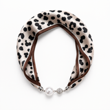 A double-loop satin neck scarf with a black cow-print pattern on a cream background, finished with a brown inner lining and a silver-tone clasp featuring a white pearl and a crystal-encrusted bead.