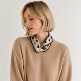 A woman with a short blonde bob wearing a beige knit sweater and a cream-colored collar scarf with black polka dots and a pearl accent, photographed against a plain light background.