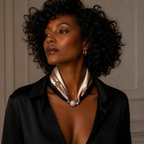 A woman with curly black hair wearing a black satin shirt and a cream silk neck scarf with a silver-toned chain and central round charm, photographed against a neutral paneled wall.