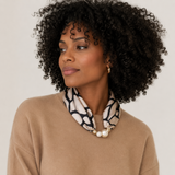 A woman with curly black hair wearing a beige knit sweater and a black and white patterned neck scarf with pearl-like beads and gold-tone hardware, photographed against a plain light background.