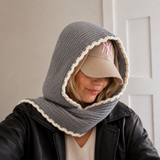 A woman wearing a gray knit hooded scarf with a cream scalloped trim, styled over a black leather jacket and a beige baseball cap with a pink NY logo.