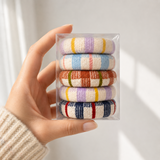 A hand wearing a beige knit sweater holds a clear plastic box containing five small striped knit hair ties or scrunchies in pastel and earthy colors against a soft white background.
