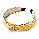 A wide fabric headband with a twisted front detail, featuring a mustard yellow and cream leaf pattern with brown beaded trim, shown on a white background.