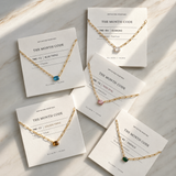 Gold necklaces with gemstones on white cards with 'The Month Code' branding on a marble surface.