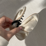 A hand holding a cream-colored fabric hair claw clip with a black plastic spring and gold-colored stitching, set against a textured off-white background with soft shadows.