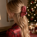 A woman with long blonde hair is shown from the back, wearing a dusty rose satin bow hair accessory with a black center. She is wearing a red plaid shirt, and a decorated Christmas tree with warm lights is blurred in the background.