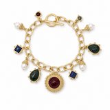 A gold charm bracelet featuring a toggle clasp and multiple dangling charms, including dark green teardrop cabochons, square blue stones, white pearl beads, and a large central round dark red cabochon, all set on a clean white background.