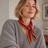A woman wearing a gray-colored knit sweater and a red knitted scarf with a mustard yellow edge, posing in a neutral indoor setting.
