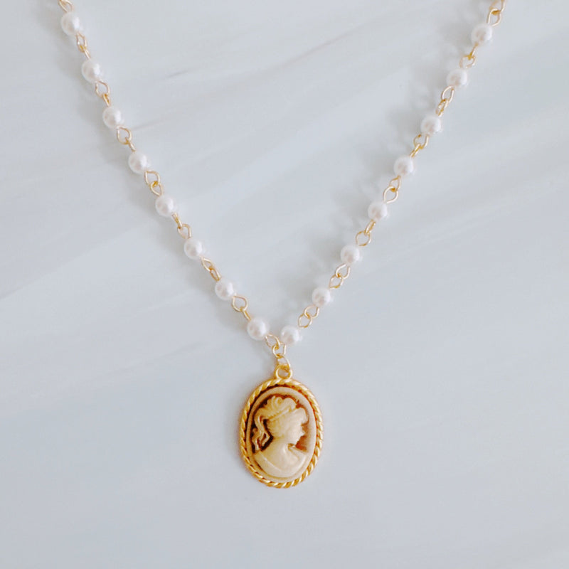 Vintage-style necklace featuring a classic cameo pendant framed in gold-tone detailing, suspended from a pearl-accented chain for an elegant and timeless look.