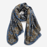 A lightweight blue scarf with an intricate paisley and floral pattern in gold, beige, and gray, draped elegantly on a light gray background.