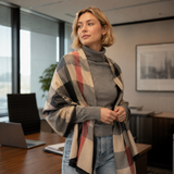A woman in a modern office setting wearing a gray turtleneck sweater and light blue jeans, wrapped in a beige plaid shawl with black and muted red stripes.