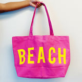 Pink tote bag with 'BEACH' in yellow letters held by a hand against a light background