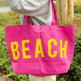 Pink tote bag with 'BEACH' in yellow letters held by a person outdoors.