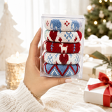 A hand holds a clear plastic box containing five pairs of folded knit socks with festive Christmas patterns in red, blue, and white. The background is a softly blurred holiday scene with a Christmas tree, wrapped gifts, and a candle.