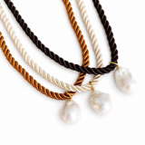 Three twisted rope necklaces in black, brown, and cream, each with a small gold ring and a baroque pearl pendant, arranged on a white background.